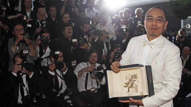 Ein Mann im weißen Anzug hält die Goldene Palme von Cannes in der Hand, umringt von Fotografen.