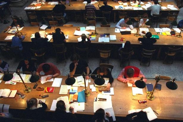 Viele Studenten sitzen an langen Tischen in einer Bibliothek und lernen.