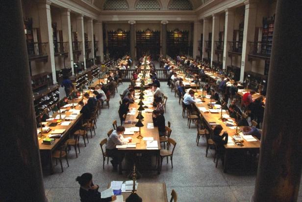 Viele Menschen sitzen an langen Tischen in einem großen Lesesaal mit hohen Bücherregalen.