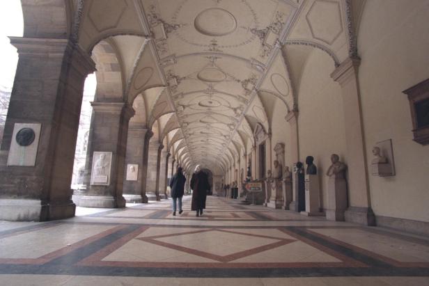 Zwei Personen gehen durch einen langen, gewölbten Gang mit Statuen und Porträts.