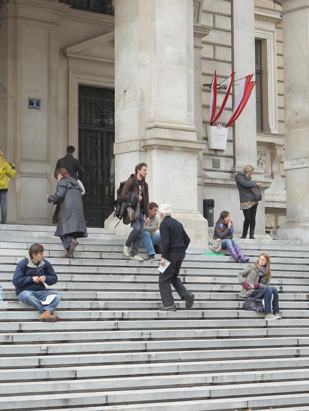 Menschen sitzen und gehen auf den Stufen vor einem Gebäude der Universität Wien.