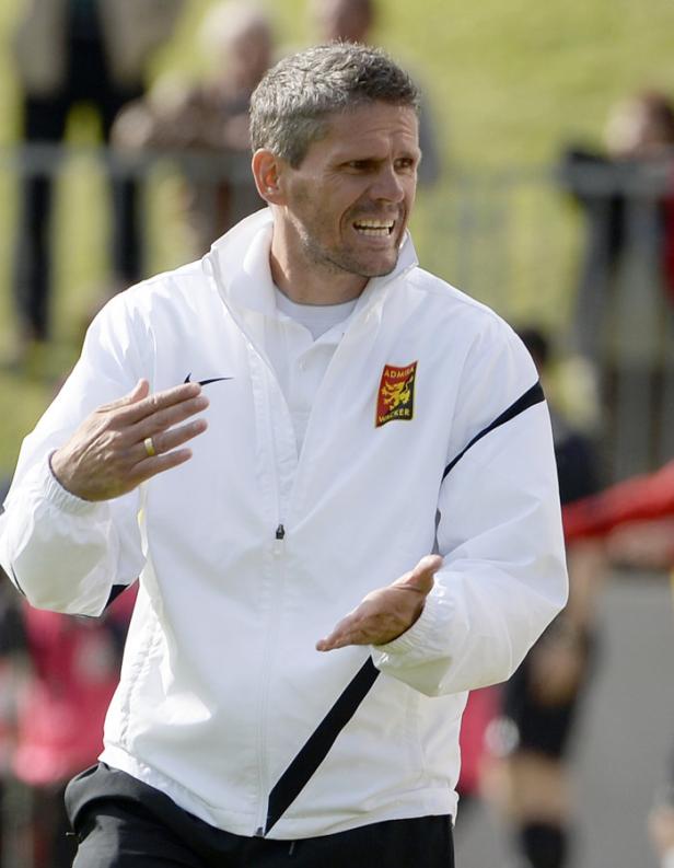 Ein Mann in einer weißen Jacke gestikuliert auf einem Fußballfeld.