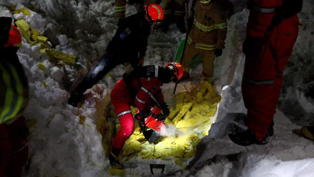 Rettungskräfte arbeiten im Schnee mit einer Kettensäge an einem gelben Objekt.