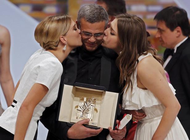 Ein Mann wird von zwei Frauen auf einer Preisverleihung links und rechts auf die Wange geküsst und hält eine Goldene Palme.