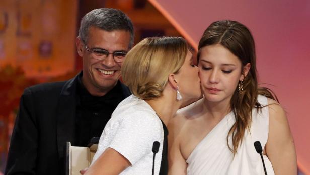 Eine Frau küsst eine junge Frau auf die Wange, während ein Mann lächelt.