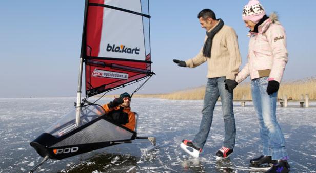 Ein Mann fährt mit einem Eis-Segelwagen auf einem zugefrorenen See, während ein Paar Schlittschuh läuft.