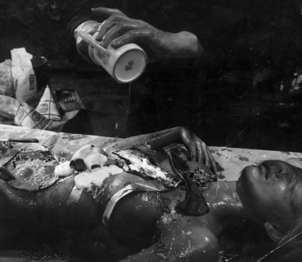 Eine Frau liegt auf einem Tisch, während eine unbekannte Substanz über ihren Körper gegossen wird.