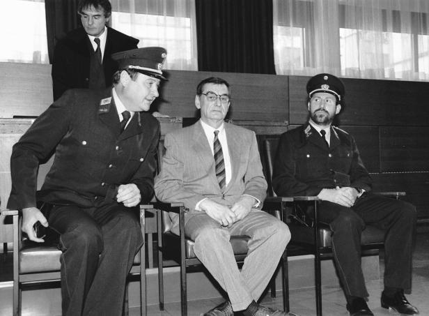 Drei Männer sitzen in Anzügen und Uniformen auf Stühlen.