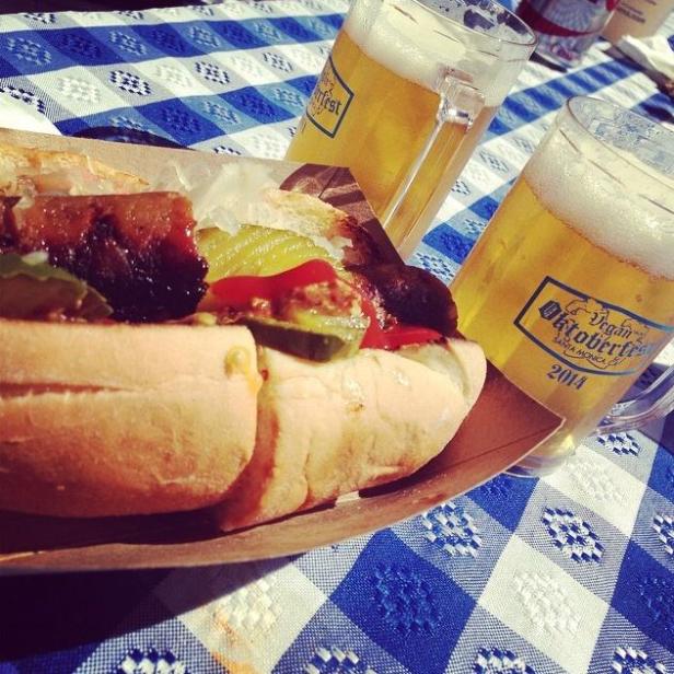 Ein Hotdog mit Gurken und Ketchup neben zwei Krügen Bier auf einer blau-weiß karierten Tischdecke.