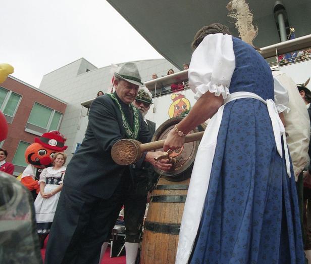 Ein Mann im Anzug schlägt ein Bierfass an, während eine Frau im Dirndl assistiert.