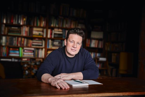 Jamie Oliver sitzt an einem Tisch mit Notizbuch vor einem Bücherregal.