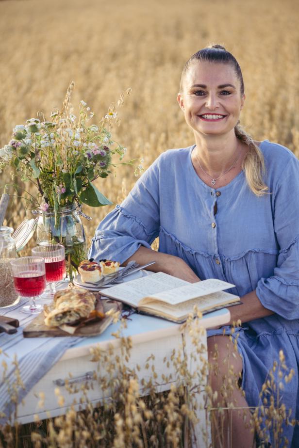 Eine lächelnde Frau sitzt an einem Tisch mit Essen und einem Buch in einem Getreidefeld.
