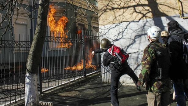 Eine Person wirft einen Gegenstand auf ein brennendes Gebäude hinter einem Zaun.