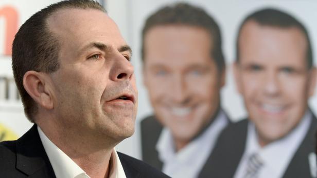 Ein Mann im Anzug spricht vor einem verschwommenen Hintergrund mit zwei weiteren Männern.