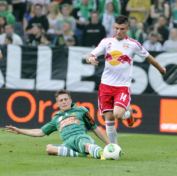 Ein Fußballspieler grätscht, während ein anderer mit der Nummer 14 den Ball führt.