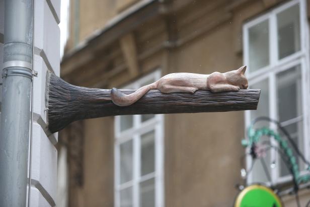 Eine hölzerne Katze liegt auf einem Ast an einer Hauswand.