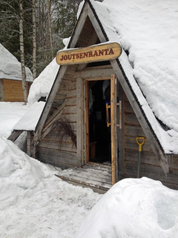 Eine schneebedeckte Holzhütte mit dem Schild „Joutsenranta“ über der Tür.