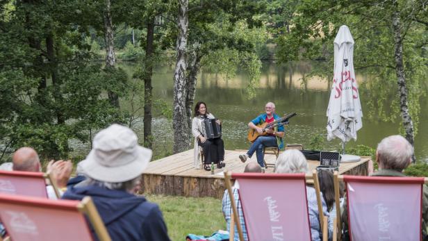Zwei Musiker spielen Akkordeon und Gitarre vor einem Publikum am See.