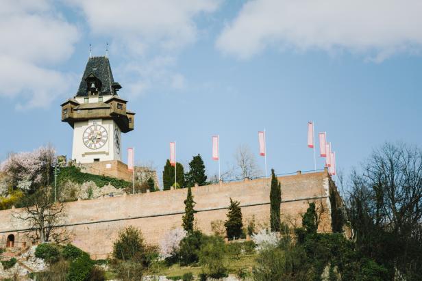 Der Grazer Uhrturm auf einem Hügel mit einer Ziegelmauer und Flaggen im Frühling.