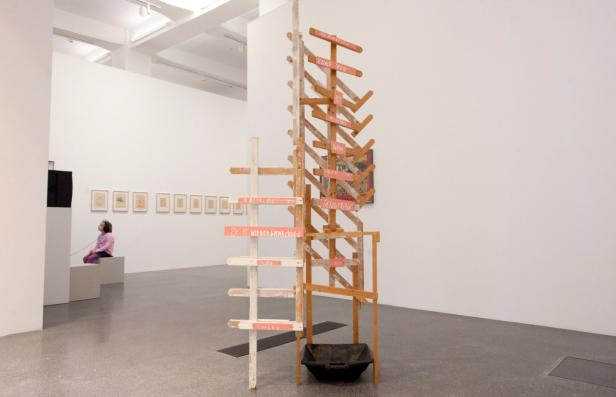 In einem Museum steht eine Holzskulptur mit Schildern, während ein Mädchen eine Videoinstallation betrachtet.