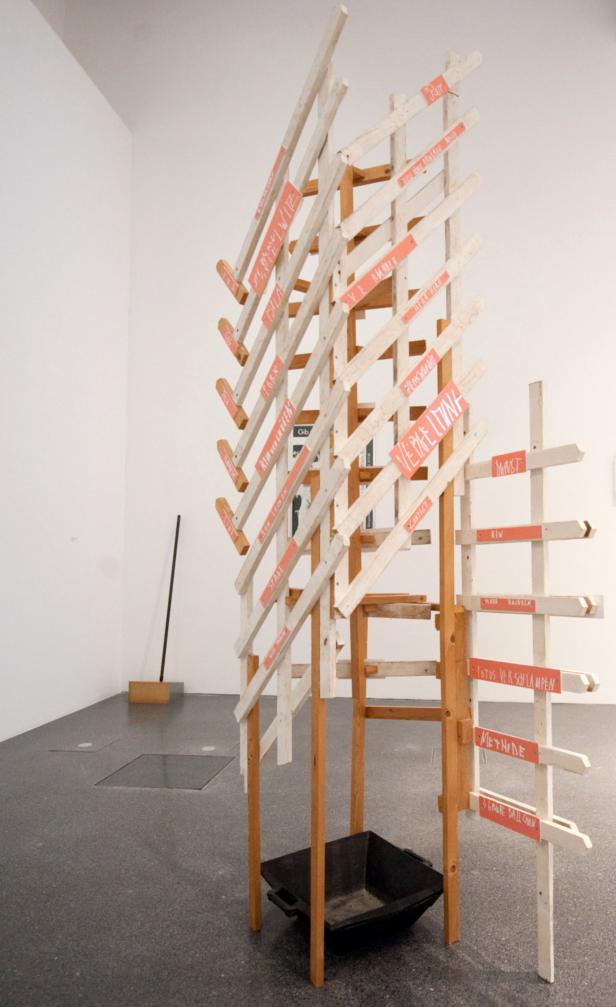 Eine Holzkonstruktion mit Schildern, die Wörter wie „Vergeltung“ und „Methode“ tragen, steht in einem Ausstellungsraum.