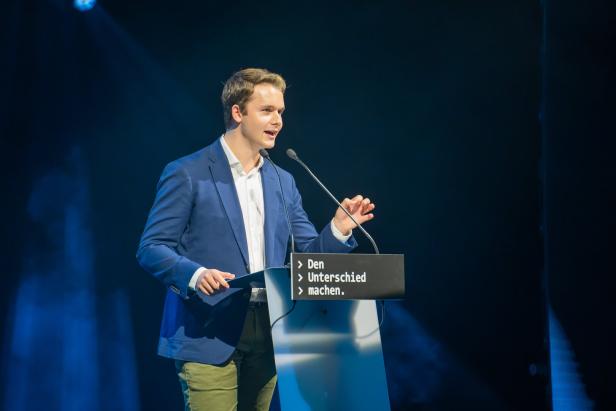 Ein junger Mann hält eine Rede auf einer Bühne vor dunklem Hintergrund.