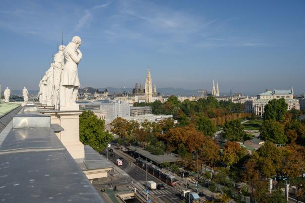 Blick über Wien mit Rathaus, Parks und Straßenbahnen von einem Dach mit Statuen aus.