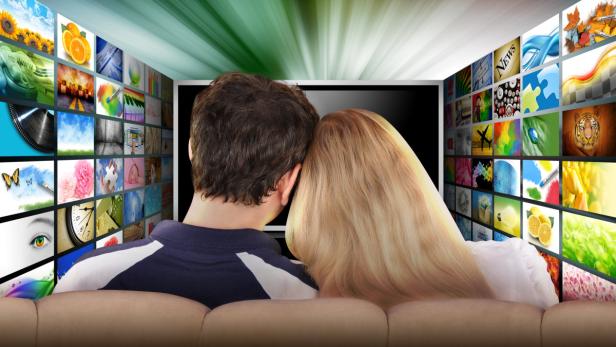 Ein Paar sitzt vor einem Bildschirm, umgeben von vielen kleinen Bildschirmen.