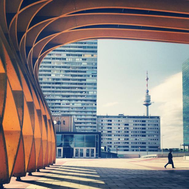 Ein Mann geht auf einem Platz in Wien vor modernen Gebäuden und dem Donauturm entlang.