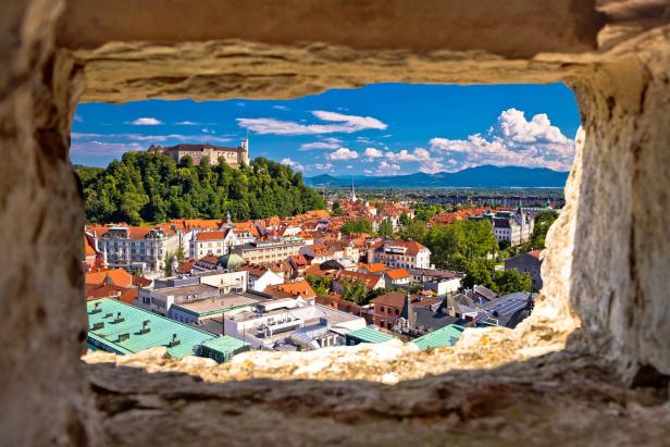 Blick durch eine Maueröffnung auf die Stadt Ljubljana mit Burg im Hintergrund.