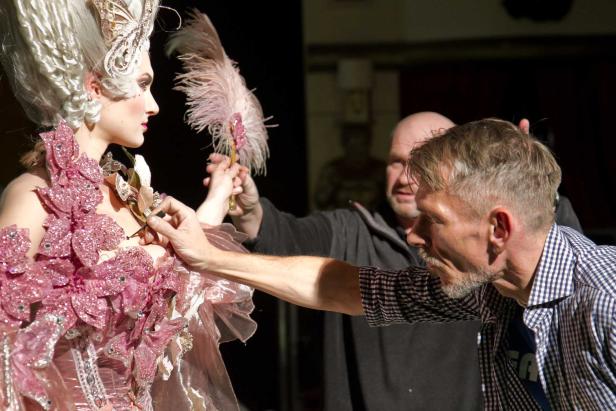 Eine Frau in einem aufwendigen Kostüm wird von zwei Personen zurechtgemacht.