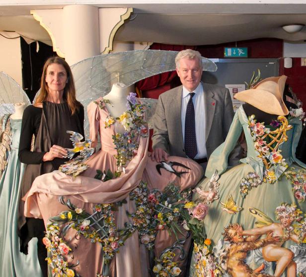 Zwei Personen posieren neben aufwändig verzierten Kleidern mit Blumen, Vögeln und mythologischen Figuren.