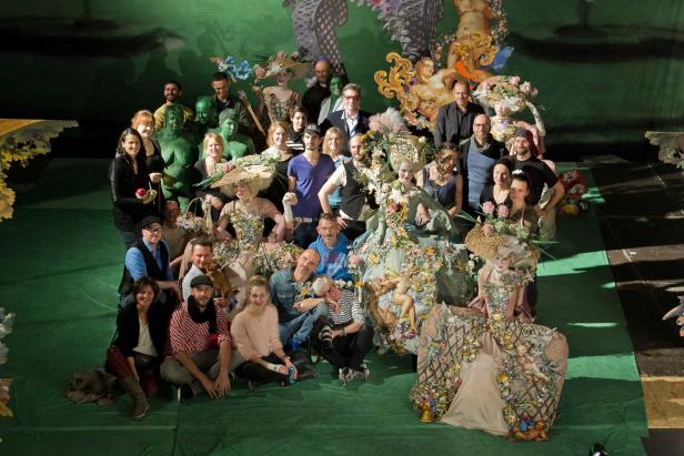 Eine Gruppe von Menschen posiert vor einer aufwendigen Bühnenkulisse mit floralen Kostümen.