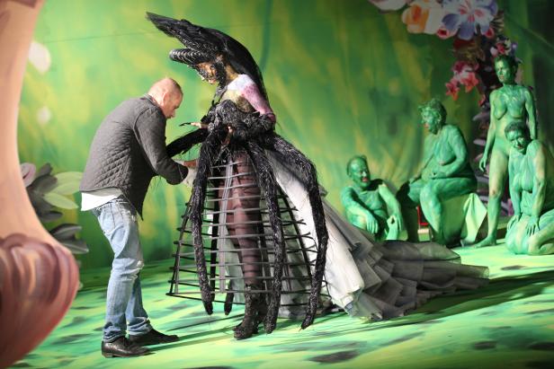 Ein Mann richtet das Kostüm einer Darstellerin auf einer Bühne, im Hintergrund grün bemalte Statisten.