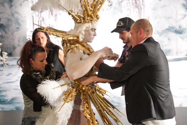 Ein Performer in einem goldenen Kostüm wird von mehreren Personen unterstützt.