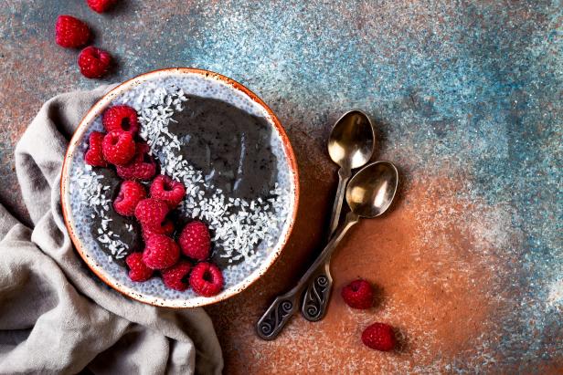 Eine Schüssel mit dunklem Smoothie, garniert mit Himbeeren und Kokosraspeln, neben zwei Löffeln.