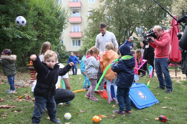 Eine Gruppe Kinder spielt mit Bällen und Spielzeug auf einer Wiese, während ein Kameramann filmt.