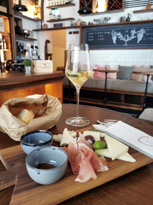 Ein Glas Weißwein und eine Platte mit Käse, Schinken und Brot in einem Restaurant.