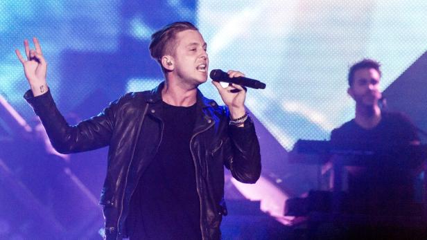 Ein Sänger mit Lederjacke performt auf einer Bühne mit blauem Licht.