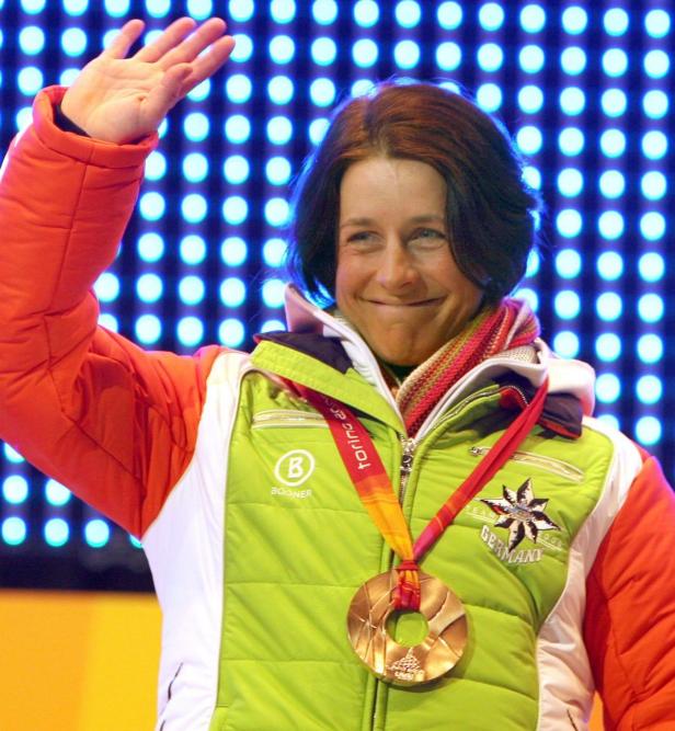 Eine deutsche Athletin mit Goldmedaille winkt vor einem blauen, punktförmigen Hintergrund.