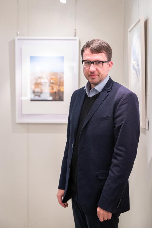 Ein Mann mit Brille und dunklem Sakko steht vor gerahmten Bildern in einer Galerie.