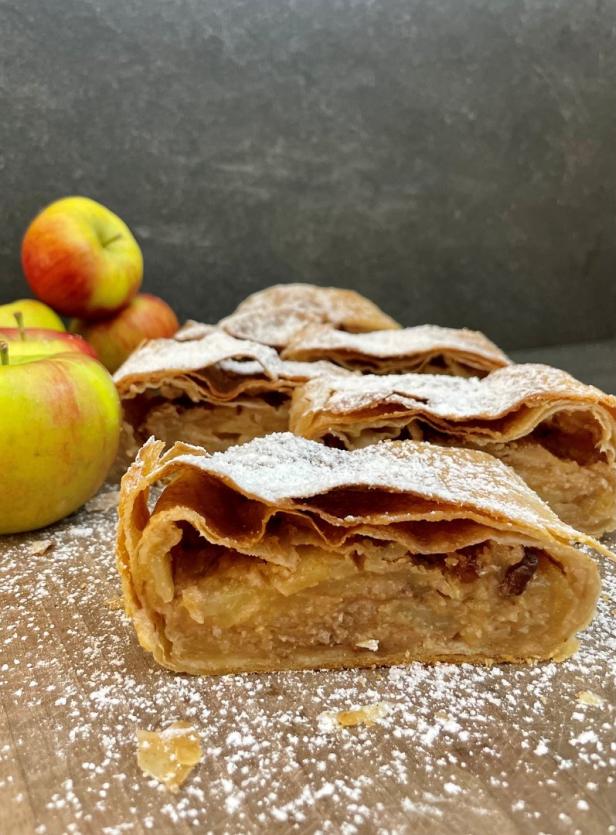 Mehrere Stücke Apfelstrudel mit Puderzucker bestreut, neben Äpfeln.