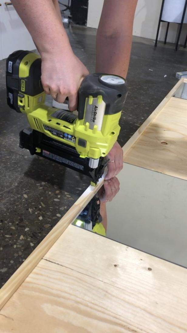 Eine Person befestigt eine Holzleiste mit einem gelben Druckluftnagler an einem Spiegel.