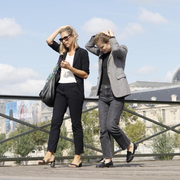 Zwei Frauen gehen auf einer Brücke in Paris und halten sich die Haare fest.