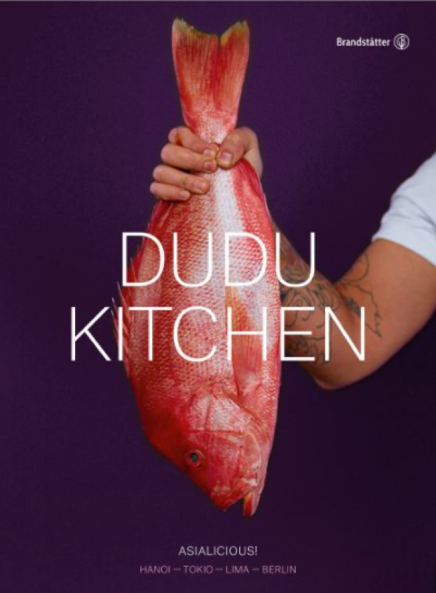 Ein roter Fisch wird vor einem violetten Hintergrund gehalten, mit dem Schriftzug „Dudu Kitchen“.