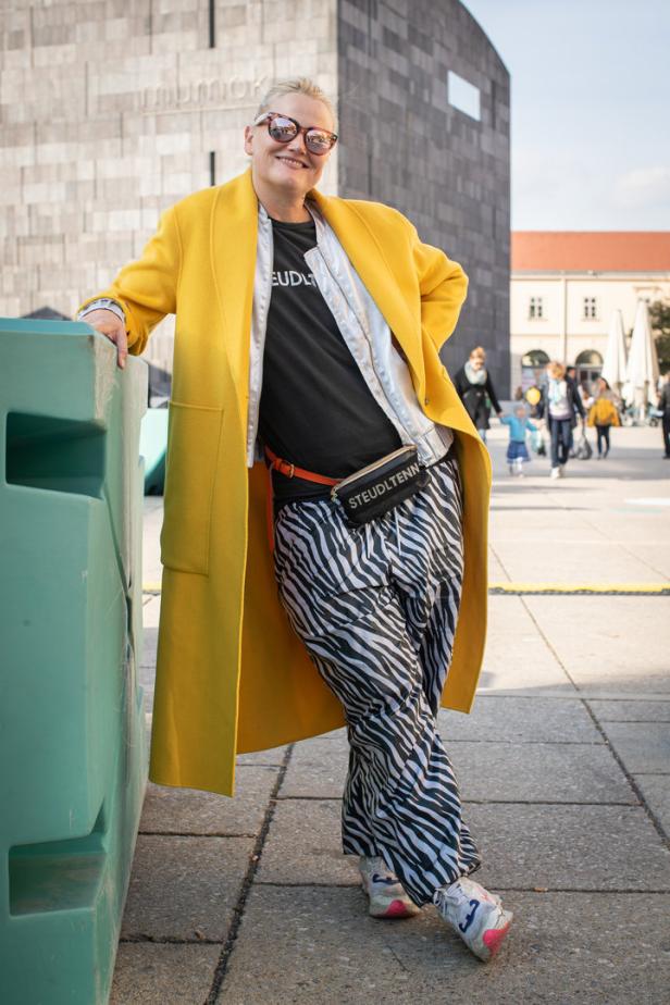 Eine Frau mit gelbem Mantel und Zebrahose posiert vor dem mumok in Wien.