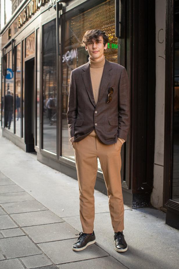Ein junger Mann in einem braunen Sakko und beigen Hose steht vor einem Louis Vuitton Geschäft.