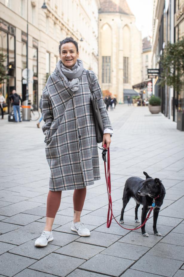 Eine Frau mit einem karierten Mantel und einem Schal geht mit ihrem Hund in einer Stadt spazieren.