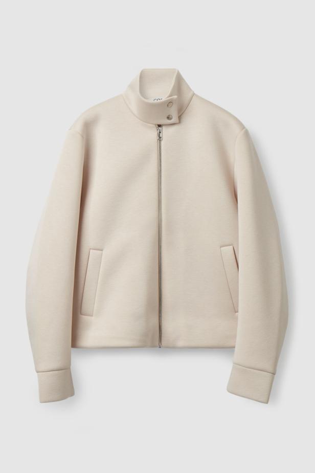 Eine hellbeige Jacke mit Reißverschluss und Stehkragen vor einem weißen Hintergrund.