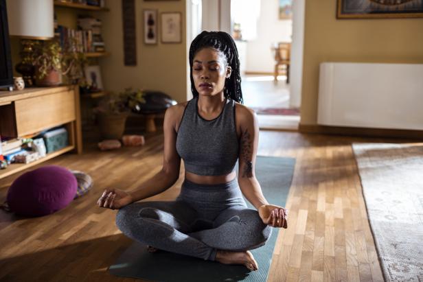 Eine Frau meditiert in einem Wohnzimmer auf einer Yogamatte.